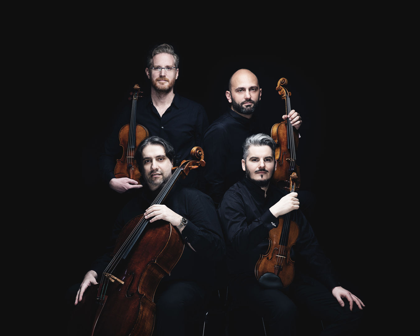 Against a black background, two men sit in front of two other men, who stand behind. All four wear black concert attire, and each man holds an instrument — clockwise from top left, violin, viola, violin, cello.